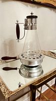 Photo showing the full coffee pot resting on the matching metal warmer with perforations, placed on a reflective surface