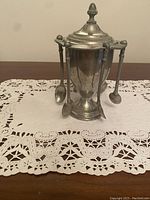 Front view of vintage silver-tone sugar bowl with four small spoons hanging on hooks attached to the bowl sides, standing on a lace doily.