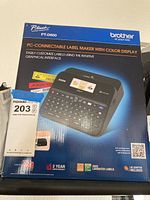 Blue product box for Brother P-touch PT-D600 label maker showing front image and model information. Indicates PC-connectable, color display, laminated label use, AC adapter included, 2-year warranty, and graphical interface.