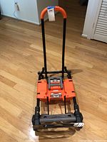Folding dolly/cart folded upright showing orange handle and folded platform on wooden floor.