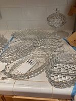 Clear pressed glass candy jar with lid and various oval and compartment serving dishes arranged on a white tile counter, showing diamond/checkered texture pattern