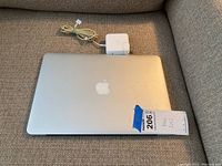 Closed Apple MacBook 2012 showing top aluminum lid with Apple logo, placed on couch alongside power adapter.