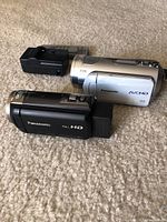 Two Panasonic video cameras (silver HC-V550 and black HDC SD1PP) side by side, with rechargeable battery on carpeted floor.