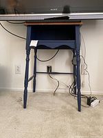 Small navy blue wooden table with natural wood top placed under a Sony TV, showing its front and side profile with visible turned legs and an open rectangular shelf below the tabletop.