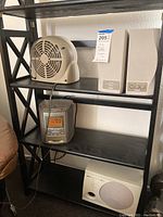 Beige electric fan, pair of beige monitor speakers, and Sony radio/CD player with digital clock display on black shelving unit.