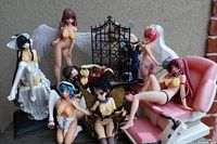 Seven anime-style figurines displayed with pink and white adjustable recliner chair and black decorative gate on brick background.