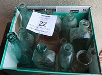 Box containing multiple antique and vintage glass bottles of various sizes and shapes, primarily pale blue-green and clear glass.