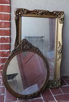 Rectangular and round antique gold mirrors leaning against a wall, showing ornate decorative gold frames and some signs of wear.