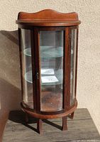 Front view of bowed half-moon glass front curio cabinet standing outside in sunlight, showing wooden frame, curved glass panes, and door.