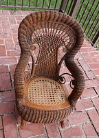 Front angle view of the child's vintage wicker rocking chair placed on a brick patio, showing the curved armrests, woven wicker backrest, and the caned seat with some wear.