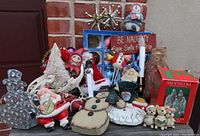 Group of assorted vintage Christmas decorations including blow mold Santa, snowmen figures, plush dolls, Shiny Brite ornament box and others on brick surface against brick wall.