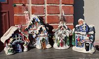 Four ceramic Christmas houses displayed outdoors on a wooden surface against a brick background, showcasing detailed colorful holiday designs and snow accents.