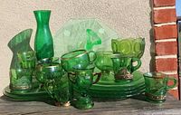 Front view of green Depression glass assortment including trays, cups with metal holders, a vase, plates, and a bubble pattern pedestal bowl. Displayed outdoors showing light reflection.