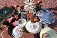 Photo showing assorted vintage ceramic plates, two Roseville vases (one pinkish pottery and one speckled), floral decorated dessert and serving plates, framed floral prints, and a round aluminum serving tray with cutouts.