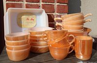 Photo of entire collection showing stackable handled mugs, handled soup bowls, ramekins, and square casserole dish with label in peach lustreware finish.