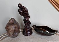 Photo showing all three items side by side on a table: a small wood-like sculpture on the left, the tall glossy brown ceramic lovers sculpture in the middle, and the brown-green glazed Beauce console bowl on the right.