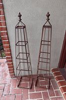 Pair of wrought iron garden obelisks standing upright on a brick floor against a wall for size and condition reference.
