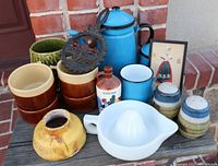 Full group of vintage kitchen items arranged on wooden surface including blue enamel coffee pot set, reamer, pottery mugs, salt and pepper shakers, vinegar bottle, green vase, cast iron trivet, and framed folk art.
