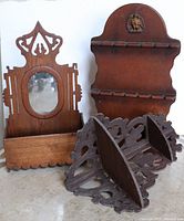 Photo showing the three wooden vintage wall-mounted items: the what-not shelf with cut-out sides and rectangular base, the spoon holder standing upright with eagle detail and pegs, and the shaving mirror with ornate sides and oval reflective glass.