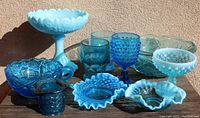Lot of vintage blue glass items on a wooden surface against a tan wall, includes hobnail goblet, trinket dishes, tumblers, pedestal compote, and bowls.