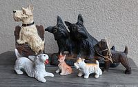 Photo of the full lot showing 7 Scottish Terrier dog items including figurines and iron doorstops in different colors and sizes on a dark wooden surface against a gray wall.
