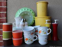 Wide view showing assortment of vintage Tupperware pieces including canisters, stackable containers, Corolle cups and saucers, salt and pepper shakers, and red condiment dispensers.