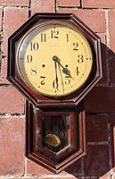 Full front view of the Hamilton vintage clock mounted on brick wall. Octagonal shape wooden frame, cream clock face with Arabic numerals, three hands showing time 5:28, glass cover protecting face.