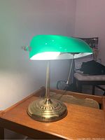 Bankers lamp on wooden table with illumination on, showing green glass shade and brass base with pull chain.