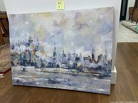 Front view of the cityscape canvas wall art on wooden frame leaning against a white wall and wooden floor.