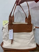 Front side of Graf Lantz travel bag showing cream canvas exterior with brown leather accent panel and handles, front zippered pocket and attached brand tag.