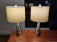 Two metal table lamps on a wooden surface, each with a brushed metal cylindrical base and beige linen cylinder shade, lit and showing the bulb.