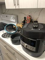Overview of the Pampered Chef electric pressure cooker on a kitchen counter with attachments including the springform pan with blue silicone ring and steaming rack.