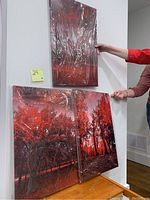 Photo showing all three canvas art pieces being held on a white wall. Each piece features red monochromatic tree landscapes with varied perspectives.