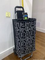 Front view of the black insulated shopping trolley with white grocery item prints. Shows the full trolley height and wheels.