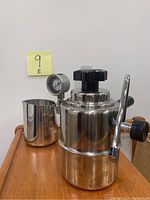 Front view of the stainless steel stovetop milk frother with attached gauge and pitcher next to it.