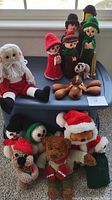 Photo showing assorted handcrafted holiday stuffed figures including Santa, snowmen, reindeer, and elves assembled on a blue plastic container and carpeted floor.