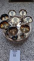 Top-down view of eight silver plated goblet cups arranged on a round, decorative metal tray. Cups show reflective surfaces with some tarnish and discoloration. Tray has a beaded edge design.
