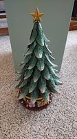 Full view of the green glittery spinning Christmas tree ornament with a gold star topper, positioned on a light-colored carpet.
