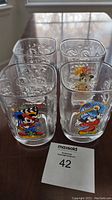 Front view of the four clear square glasses with colorful Mickey Mouse millennium 2000 prints and embossed swirl patterns, arranged on a wooden surface with a MaxSold lot card.