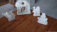 White ceramic nativity scene pieces displayed on a wooden table including stable, angel, shepherds, and sheep