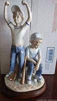 Photo showing full figurine of two boys in baseball uniforms on wooden base, one boy standing with arms raised, other sitting with bat and balls