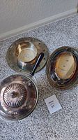 Three silver-tone serving dishes on carpeted floor; tarnished with no visible markings, one has black handle and ribbed lid.
