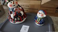Pair of ceramic snowman and snowmama candle holders shown with box, placed on gray surface.