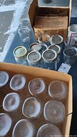 Box containing 31 assorted Ball and Kerr mason jars with various metal lids. Jars are cleanable, showing some residue and use. Some jars still in original packaging.
