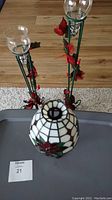 Three tea light candle holders visible on a tray; two tall green metal holders with glass globes and red artificial rose decorations, one stained glass lamp with floral design and bronze metal base.