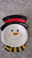 Front view of snowman shaped platter with painted black top hat with red band, smiling snowman face, orange nose, and green-yellow scarf.