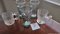 Five glasses on table: three green embossed Coca Cola glasses and two clear Flintstones 1993 mugs.