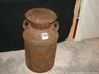 Full view of the rusty metal milk can standing upright showing the cylindrical body, top lid, and side handles.