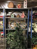 Metal shelving unit with three birdhouses on top shelf, two Easter egg wreaths on middle shelf, artificial plant and Christmas tree on bottom shelf
