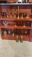 Full view of shelving unit with assortment of brass candlesticks, glass candlesticks, wooden candlesticks, metal pitchers, and perforated metal containers.
